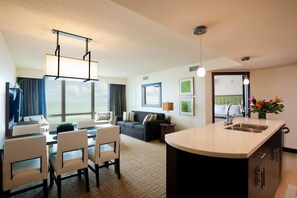 A hotel room with a dining area, a sofa, a TV, a large window, and a kitchen area.