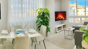 A modern living room with a dining area, a large plant, a flat-screen TV, and a view of buildings through large windows. - Cozy 1BR condo w/ King bed, full AC, WiFi, smart TVs & Full kitchen in El Vergel (Santo Domingo)