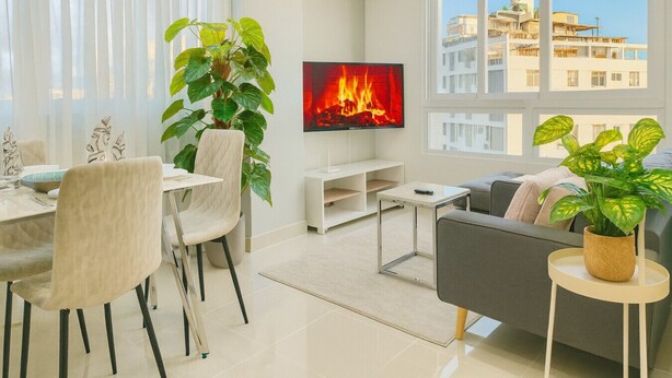 A modern living room with a flat-screen TV, a dining area with chairs and a table, and large windows offering a view of buildings. - Cozy 1BR condo w/ King bed, full AC, WiFi, smart TVs & Full kitchen in El Vergel (Santo Domingo)