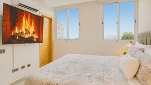 A bedroom with a large bed, a TV mounted on the wall, and a window with a view of buildings. - Cozy 1BR condo w/ King bed, full AC, WiFi, smart TVs & Full kitchen in El Vergel (Santo Domingo)