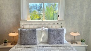 Un dormitorio con cama, dos mesitas de noche, un ventanal con vistas a palmeras y una colcha estampada.