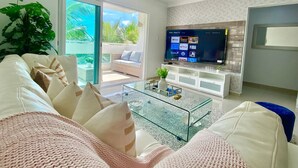 Una sala de estar moderna con una mesa de centro de vidrio, sofás blancos y un televisor de pantalla plana mostrando varios canales.