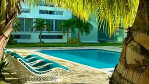 Zona de piscina con sillones de descanso, palmeras y un edificio moderno al fondo.