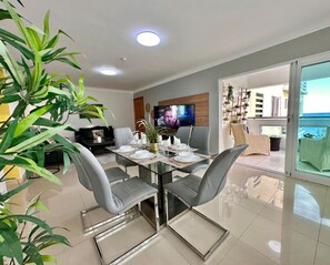 A modern dining area with a glass table, grey chairs, and a TV on the wall. - Beautiful Ocean View Apt. Pool (Santo Domingo Este)