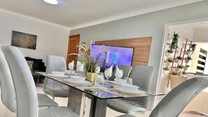 Un comedor moderno con una mesa de vidrio, sillas tapizadas de color gris y un televisor al fondo.