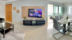 Una sala de estar moderna con un televisor de pantalla plana montado en la pared, un comedor con una mesa de vidrio y sillas, y una puerta de madera.