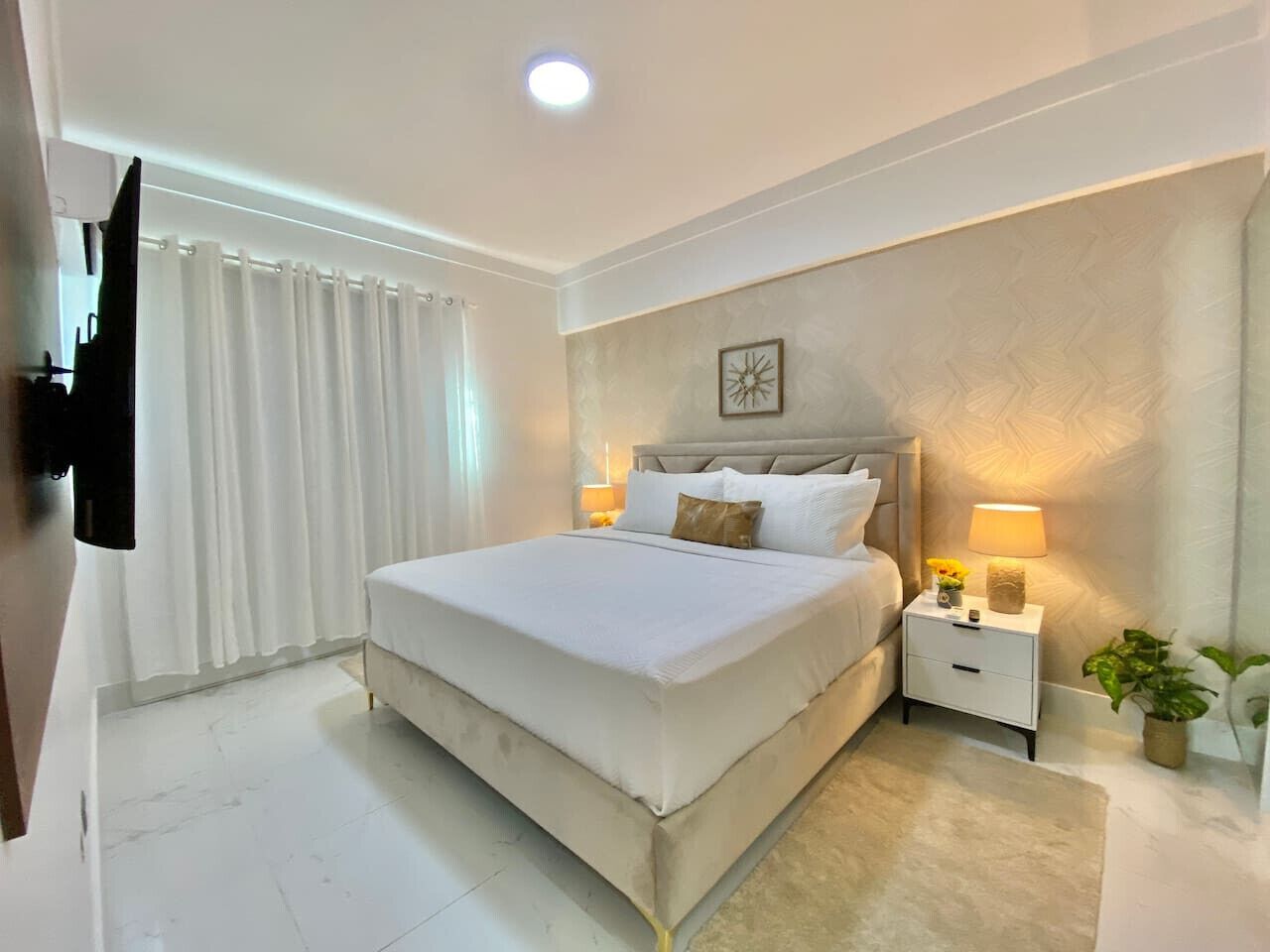 A modern bedroom with a large bed, white curtains, a nightstand with a lamp, and a wall-mounted TV.