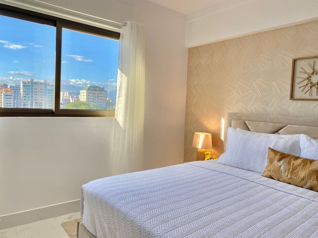 A hotel room with a large bed, a nightstand with a lamp, a window with a view of buildings, and a wall clock.