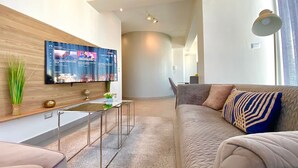 A modern living room with a flat-screen TV mounted on the wall, a sofa with patterned cushions, a glass coffee table, and a small side table with decorative items.