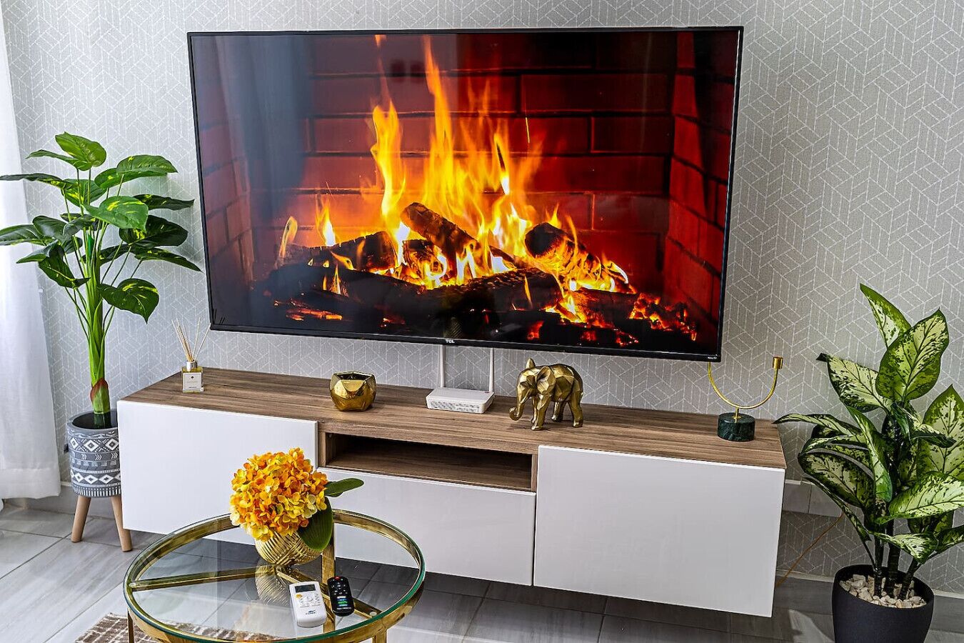 A modern living room with a flat-screen TV mounted on a wooden console, a glass coffee table with a vase of flowers, and two potted plants.