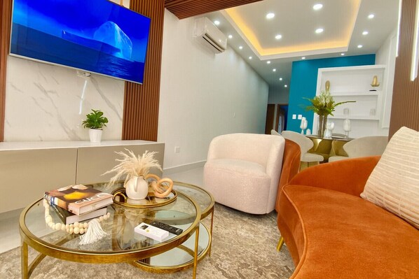 A modern living room with a glass coffee table, a white armchair, and an orange sofa. A flat-screen TV is mounted on the wall, and there is a bookshelf in the background. - Elegant, Finely Decorated City Center 2 Bdrm Condo, Pool, Near All (Santo Domingo)