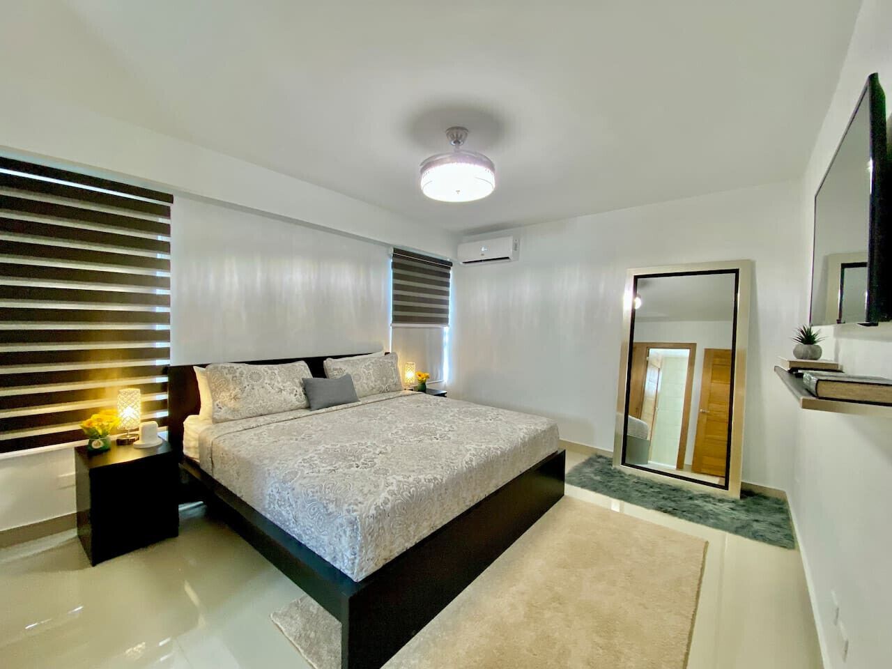 A modern bedroom with a large bed, bedside tables, a ceiling light, a mirror, and a wall-mounted air conditioner.