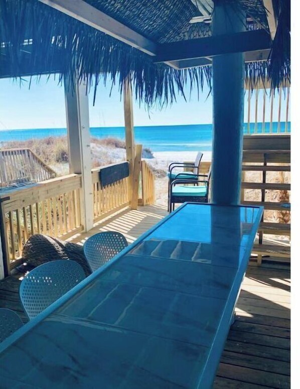 Relax in a covered outdoor seating area with a long table and comfortable chairs, overlooking the ocean and sandy beach.
