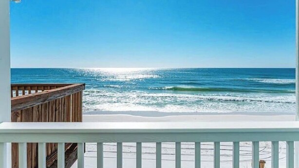A wooden deck with a white railing overlooking a beach with clear blue water and a bright sun reflecting on the surface. - Sunrise, Sunburn, Sunset, Repeat (Destin)