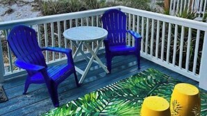 A deck with two blue chairs and a small table, overlooking a beach with sand and grass. - Sunrise, Sunburn, Sunset, Repeat (Destin)