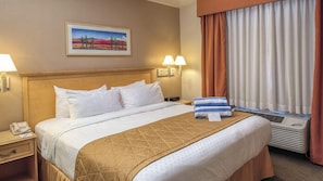 A hotel room with a bed, a nightstand, a lamp, a painting on the wall, and a window with curtains. - Tucson | Relaxed Studio Retreat | Pool + Hot Tub (Tucson)