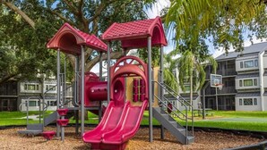 A playground with slides and climbing structures, surrounded by trees and a building in the background.