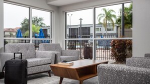 A hotel room with a grey sofa, a wooden coffee table, a suitcase, and large windows with a view of palm trees and buildings.