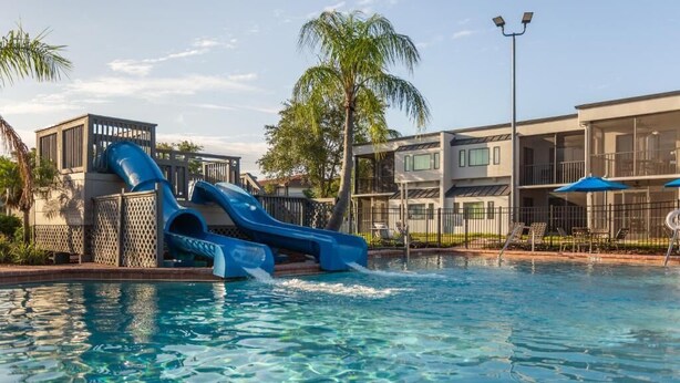 A pool with a blue water slide, palm trees, and modern buildings in the background.
