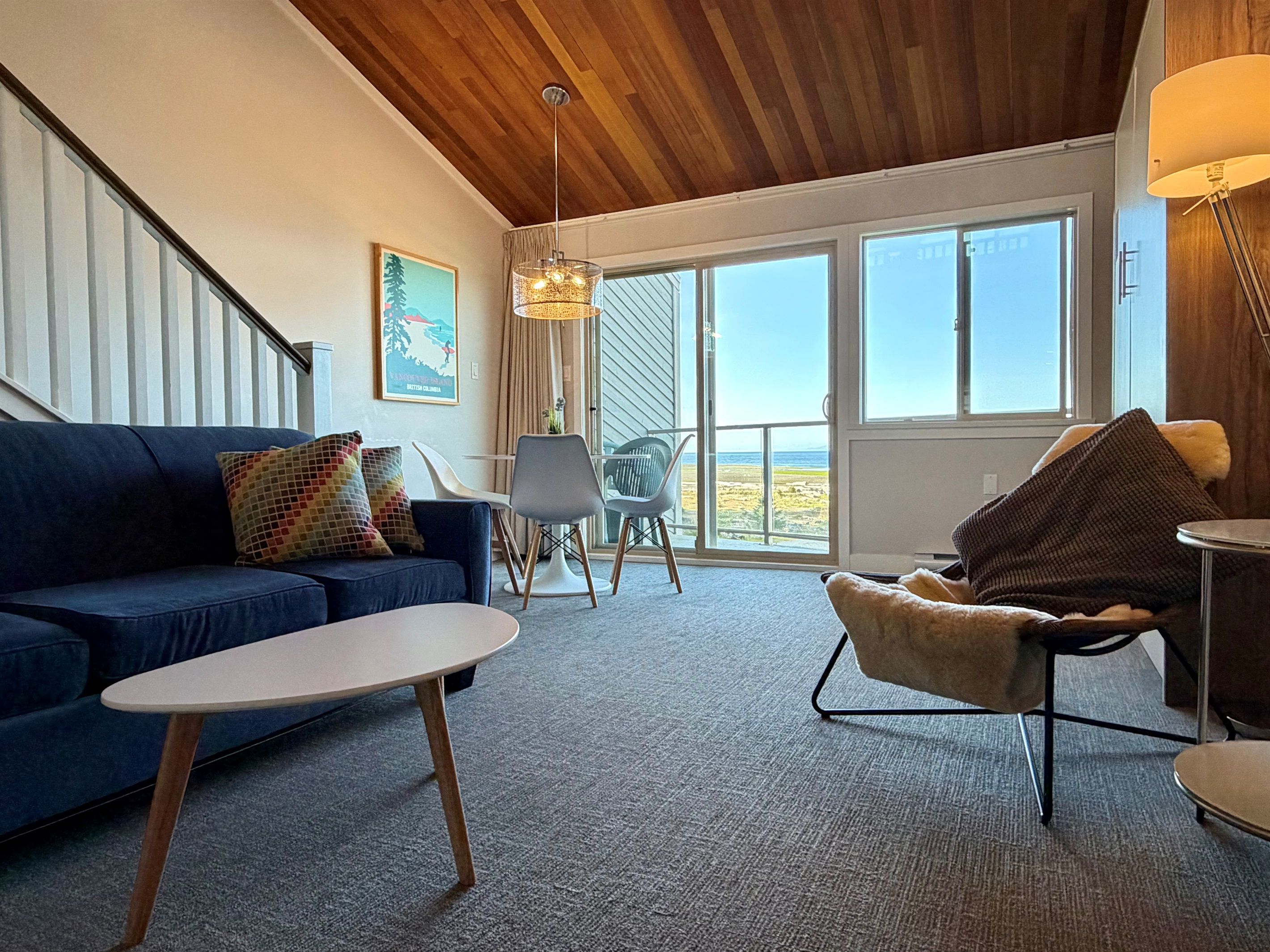 A room with a blue sofa, a small table, a chair, a lamp, and a view through large windows.