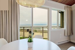 Una habitación con una mesa blanca, una planta en maceta, vista a la playa y un ventanal con cortinas.