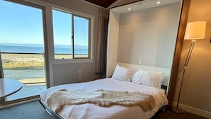 A hotel room with a bed, a round table, a lamp, and a view of the beach. - Oceanfront Studio with Loft - amazing views! (Qualicum Beach)