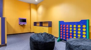 Une pièce avec un mur jaune, un meuble TV violet, un plateau de jeu bleu et jaune, et deux poufs noirs.