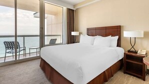 A hotel room with a large bed, a nightstand with a lamp, a chair, and a view of the sea through a sliding glass door.