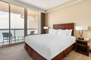 Ein Hotelzimmer mit einem großen Bett, einem Nachttisch mit Lampe, einem Sessel und Blick auf das Meer durch eine Glastür.