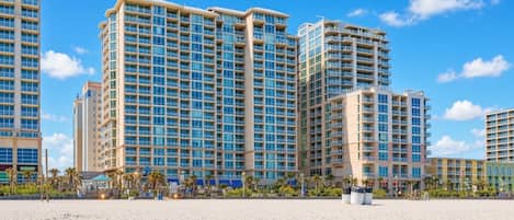 A high-rise hotel with multiple balconies, a sandy beach in the foreground, and palm trees.