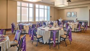 A banquet hall with round tables, chairs, and purple sashes, featuring large windows and ceiling lights.