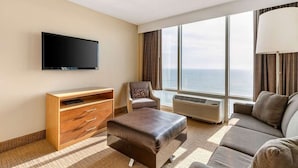 A hotel room with a flat-screen TV, a sofa, a chair, a wooden cabinet, and a large window with a view of the ocean. - Virginia Beach | Ocean Views | Pool & Beach | 1BR Suite (Virginia Beach)