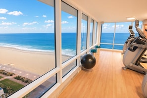 Un gimnasio con equipo de ejercicio y un amplio ventanal con vistas a la playa.