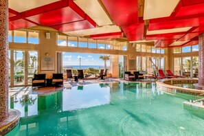 An indoor swimming pool with clear blue water, surrounded by lounge chairs and a view of the ocean.