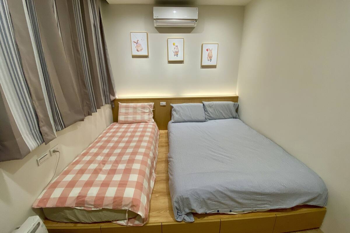 Een slaapkamer met twee eenpersoonsbedden, een raam met gordijnen, een airconditioner en drie ingelijste foto's aan de muur.