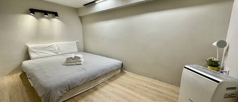 A small, clean room with a bed, a bedside table, a lamp, and a heater.