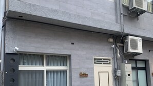 A building with a grey facade, a beige door, and a window with curtains. There are two benches outside, a trash bin, and an air conditioning unit on the wall. - Liu-He 914 Hostel (Kaohsiung)