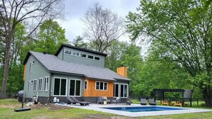 Una casa de dos pisos con un amplio patio delantero, una piscina y mobiliario de exterior.