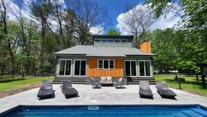 Una casa moderna de dos pisos con piscina, zona de estar exterior y vistas a los árboles circundantes.