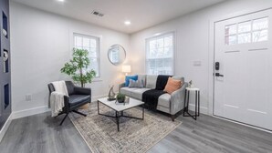 A modern living room with a grey sofa, a black armchair, a coffee table, and a small side table. There are two windows with blinds, a door, and a mirror on the wall. - Modern Uptown Getaway & Backyard Oasis (Charlotte)