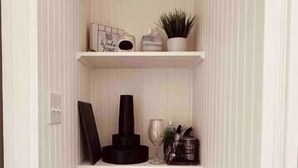 Un couloir étroit aux murs blancs et au plancher en bois. On y trouve trois étagères sur lesquelles sont disposés divers objets, notamment une plante en pot, une machine à café et une couverture enroulée.