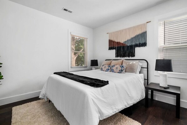 A modern bedroom with a large bed, a bedside table with a lamp, a window with blinds, and a wall-mounted tapestry.