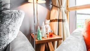 Een knus ogende kamer met een comfortabele grijze bank, een klein houten bijzettafeltje met decoratieve kerstboompjes en een rode kandelaar, een lamp, en een raam met gordijnen.