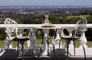Ein Balkon mit weißen schmiedeeisernen Stühlen und einem Tisch, mit Blick auf eine Stadtlandschaft.