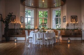 Ein Zimmer mit einem runden Tisch, gedeckt für sechs Personen, umgeben von weißen Stühlen, großen Fenstern mit Vorhängen und einer Holzbalkendecke.
