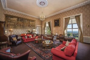 Ein geräumiges Wohnzimmer mit einem großen roten Sofa, Ledersesseln und einem Couchtisch. Es gibt eine Chandelier, ein großes Gemälde an der Wand und einen Ausblick durch das Fenster.