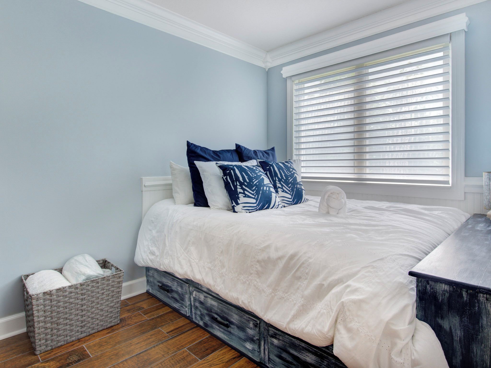 Un dormitorio con cama, ropa de cama blanca, un cesto de almacenamiento gris y una ventana con persianas.