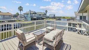 A deck with wicker furniture, a glass table, and a view of a water body and buildings in the background. - Celestial Dreams by Ryson Vacation Rentals (Galveston)