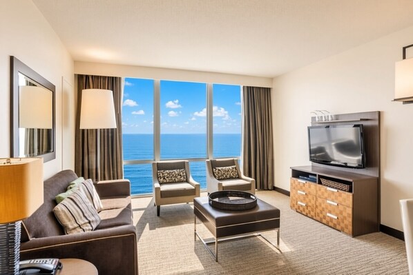 A hotel room with a sofa, armchairs, a coffee table, a TV, and a view of the ocean.