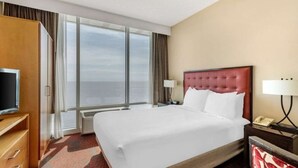Habitación de hotel con una cama grande, televisión, escritorio con lámpara y vista al océano a través de amplias ventanas.